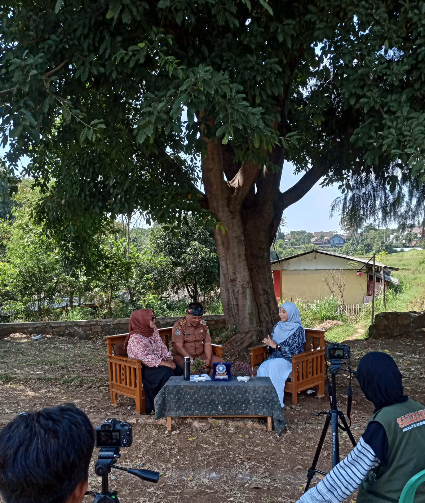 Momen KKN IPB University saat merekam program Bincang Hangat Desa, yang bertujuan untuk menggali isu dan harapan dari Kepala Desa dan Ketua PKK secara langsung. Latar alam yang asri dan suasana yang informal menunjukkan pendekatan yang personal dalam komunikasi.