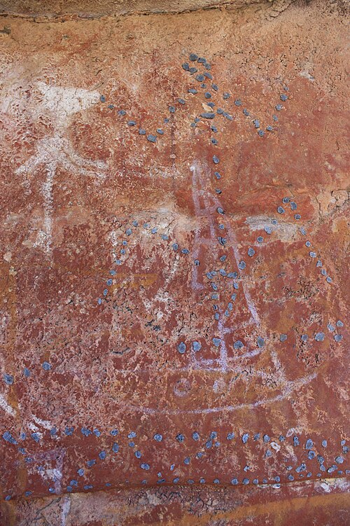 Lukisan seni cadas di Arnhem Land yang menggambarkan sebuah perahu Makassan berwarna putih, dengan figur perempuan yang digariskan menggunakan lilin lebah (beeswax) di atasnya. Teknik penggarisan beeswax di atas lukisan seperti ini adalah salah satu ciri khas seni cadas Arnhem Land, dan motif perahu Makassan menunjukkan bagaimana masyarakat Yolngu turut merekam kehadiran pelaut Nusantara dalam karya visual mereka. Foto: Wikimedia Commons (Domain Publik).