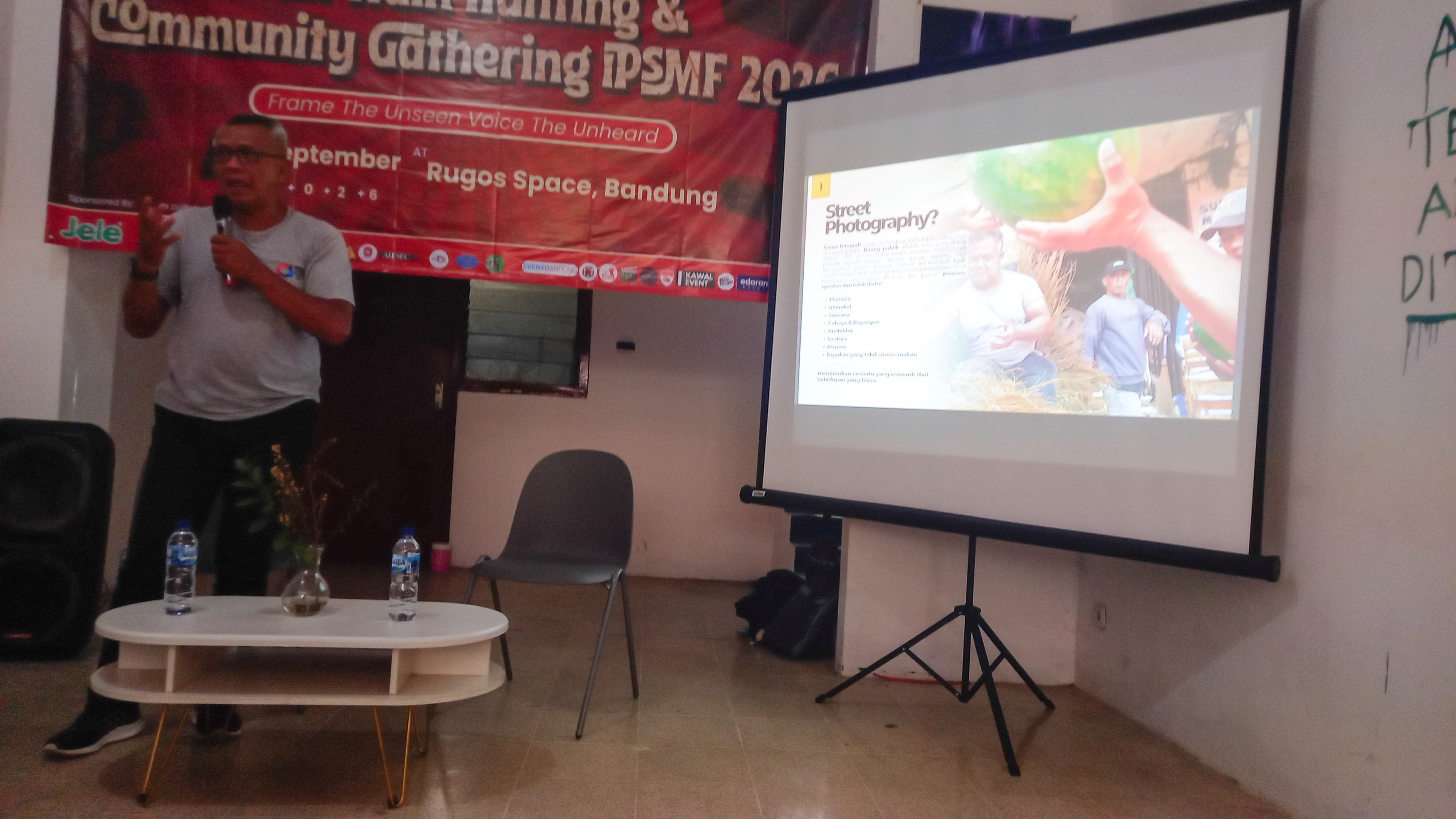 Arif Rudiana sebagai pemateri sedang berbicara memegang mikrofon di samping layar proyektor presentasi Street Photography dalam acara Kawan GNFI x IPSMF 2026 di Rugos Space Bandung