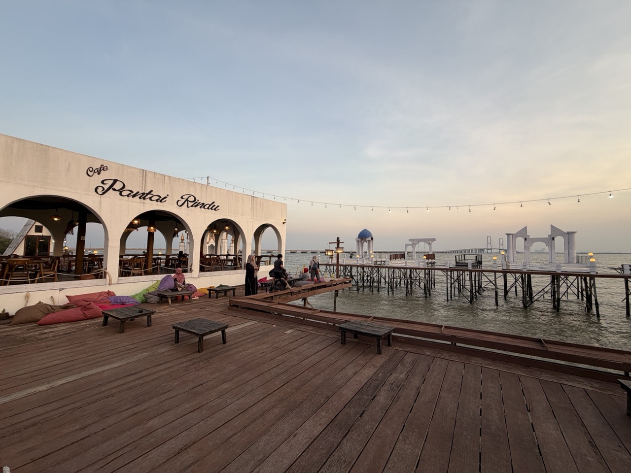 Visitors unwind at Café Pantai Rindu as dusk settles over the Madura Strait, with Suramadu Bridge visible in the distance.