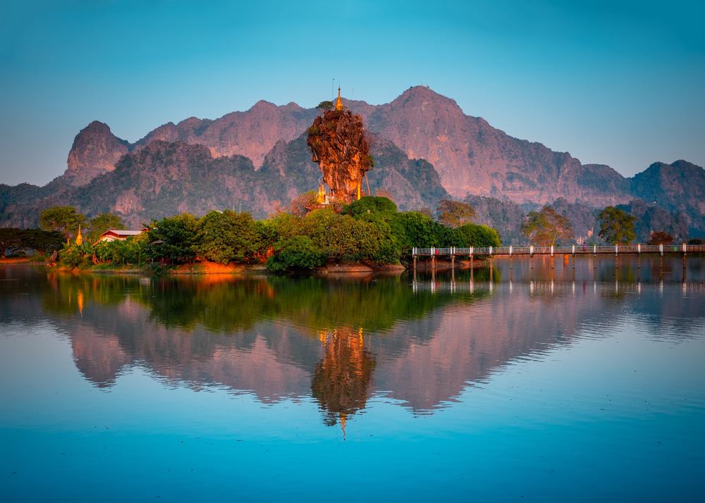 A tall limestone rock formation rising from a tranquil lake, with lush greenery and distant mountains in Hpa-An, Myanmar.
