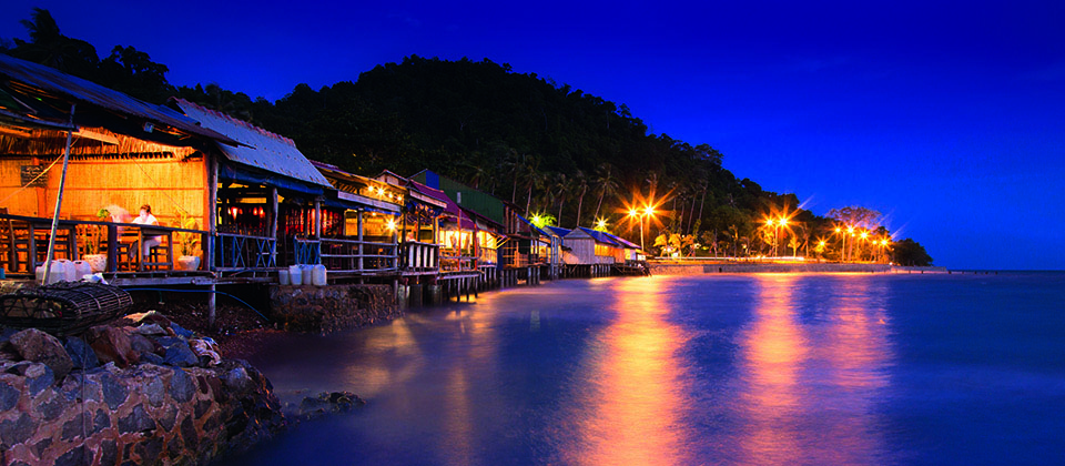 Evening view of the coastline in Kep, Cambodia, with warm lights reflecting on calm seawater.