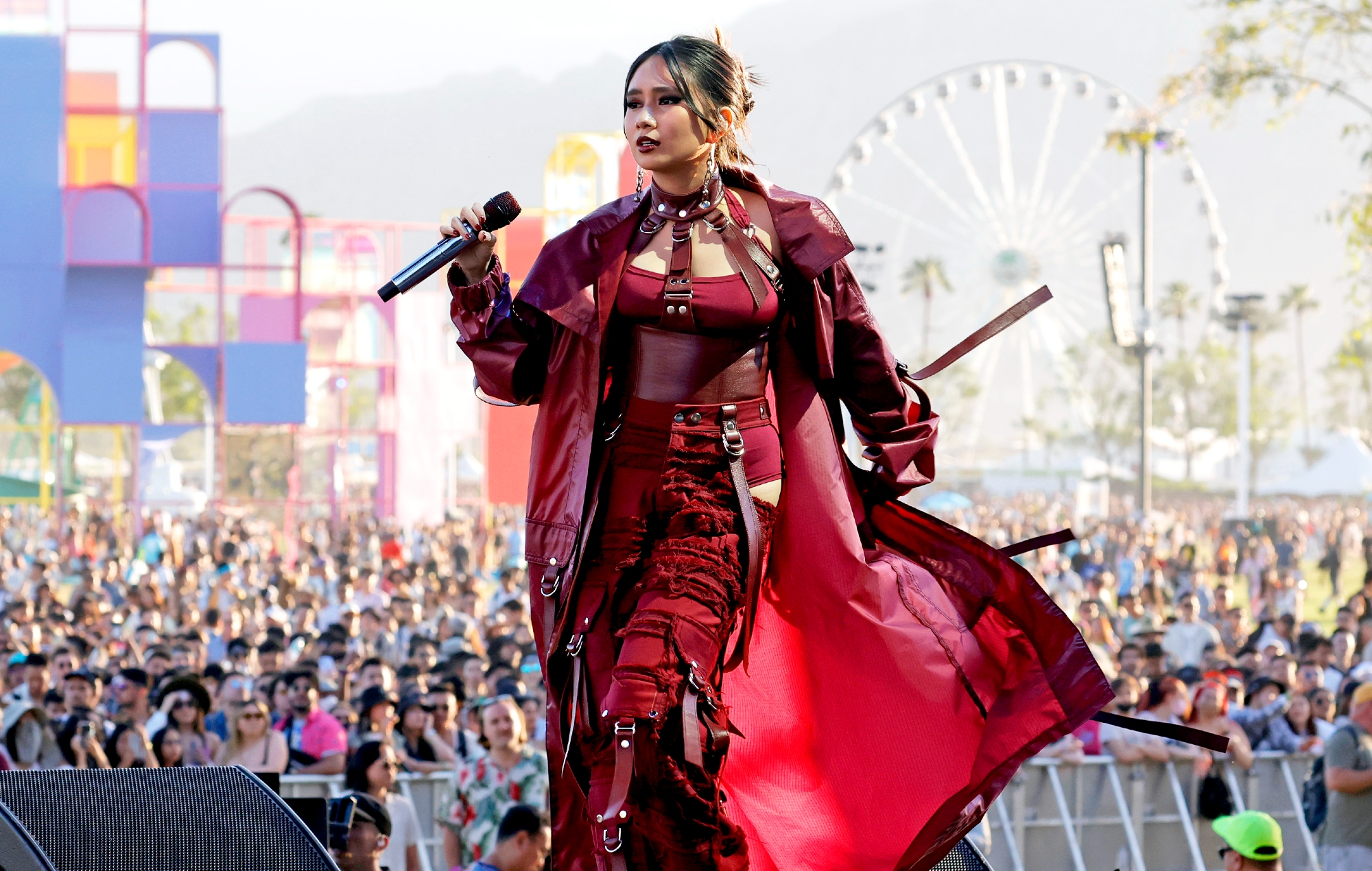 A female singer in a bold red outfit performs on stage at an outdoor music festival, with a large crowd and a Ferris wheel in the background.